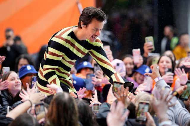 Singer Harry Styles, wearing a colourful striped jumpsuit, bends down from a stage to greet enthusiastic fans