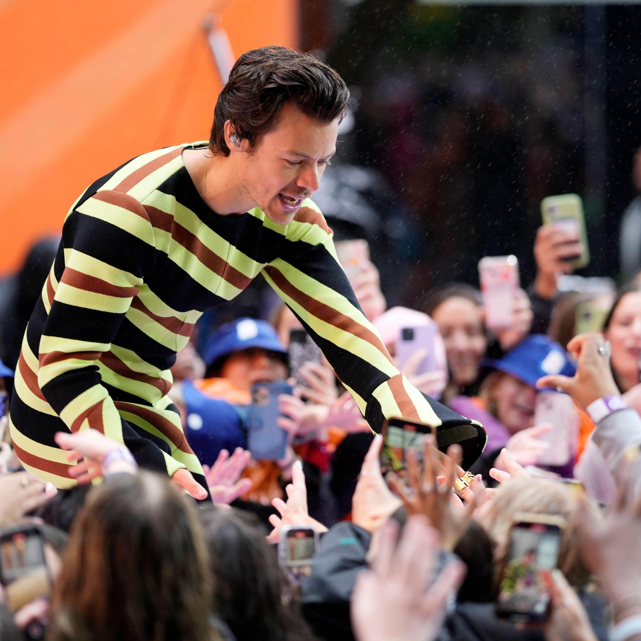 Singer Harry Styles, wearing a colourful striped jumpsuit, bends down from a stage to greet enthusiastic fans