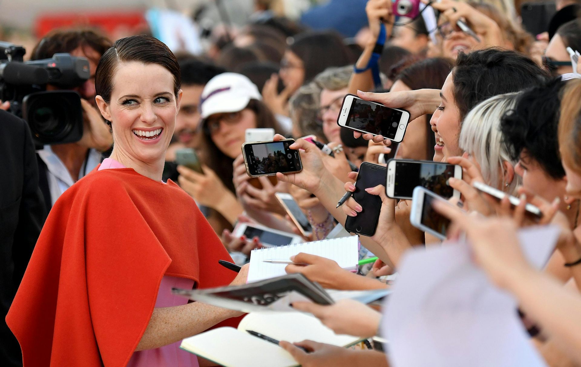 Claire Foy in a red, cape-like dress smiles over her shoulder while a crowd of fans take photos and ask for autographs