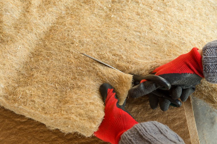 Close up shot of person with red gloves and sharp cutting tools cutting pale brown panel of hemp fibrous insulation material