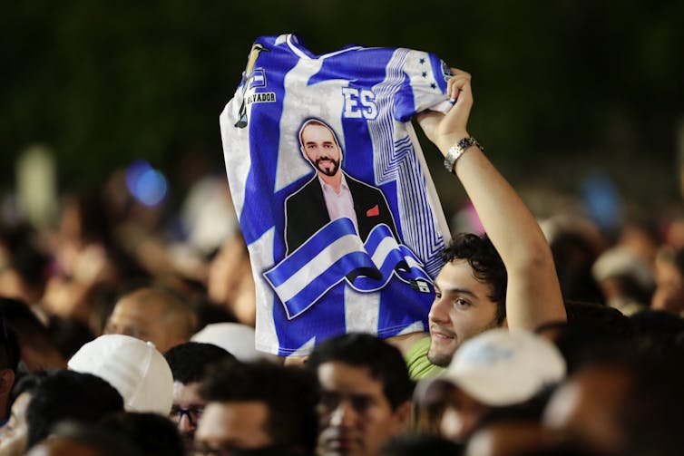 Un hombre en una multitud sostiene en alto una camiseta con la imagen de Nayib Bukele impresa en el frente.