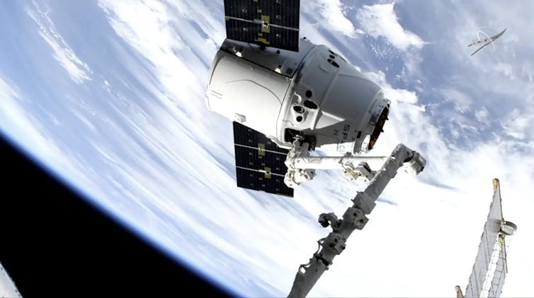 Biały statek kosmiczny w kształcie stożka z dwoma prostokątnymi panelami słonecznymi w przestrzeni kosmicznej, z Ziemią w tle.