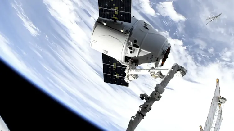 A conical white spacecraft with two rectangular solar panels in space, with the Earth in the background.