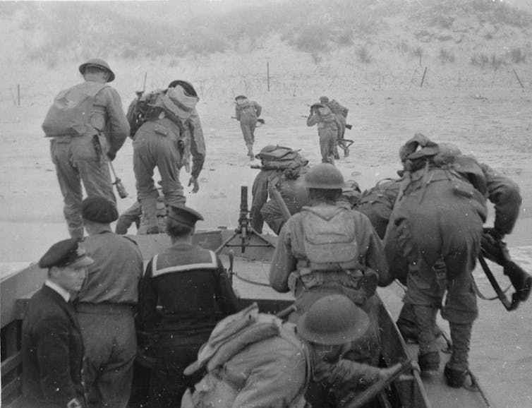 People wearing military uniforms bend forward and advance through sandy terrain.