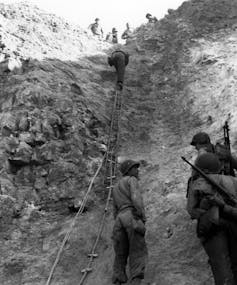 Soldiers stand below ladders rising up a cliff, where other soldiers stand.