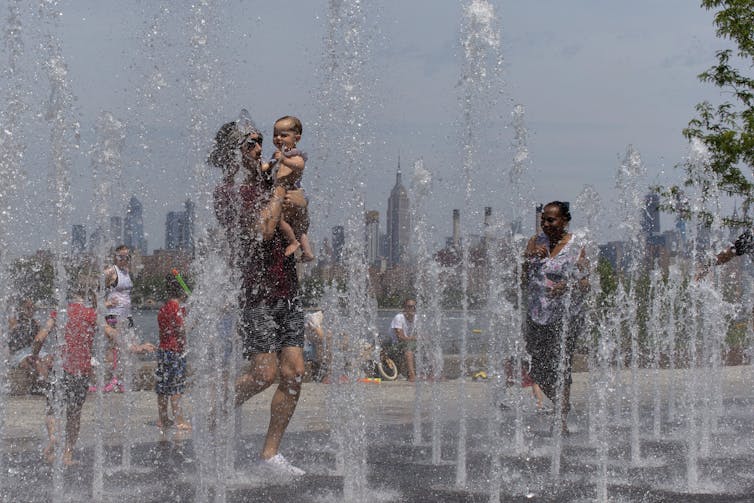 A man carries a baby in a fountain with spray coming from the ground on a hot day.