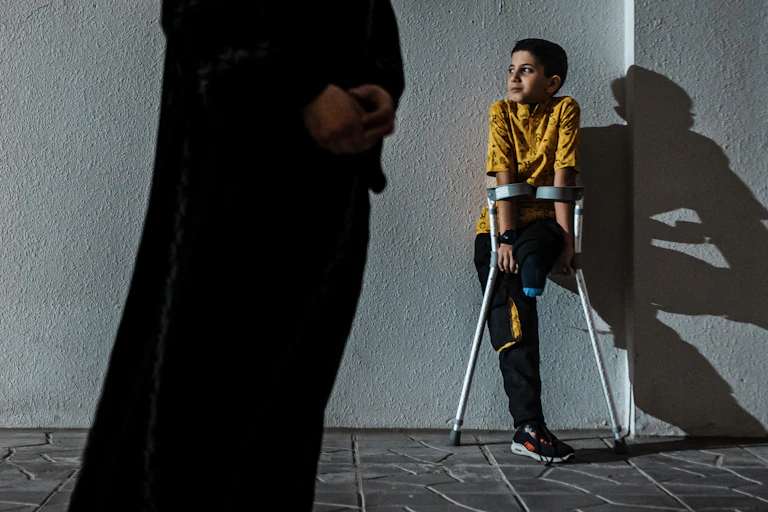 Young boy without a leg uses crutches as he leans against a wall. A faceless adult stands in the foreground.