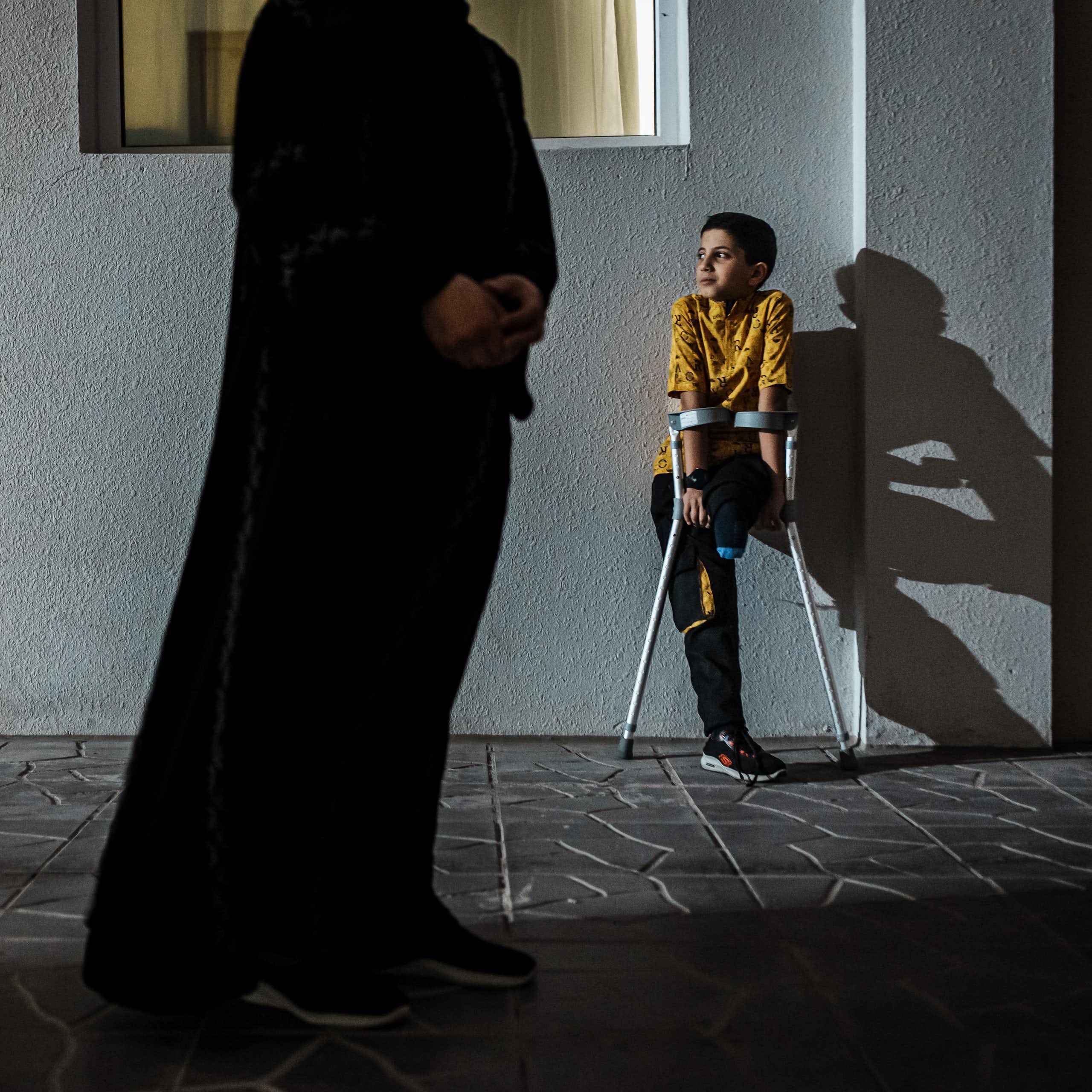 Young boy without a leg uses crutches as he leans against a wall. A faceless adult stands in the foreground.