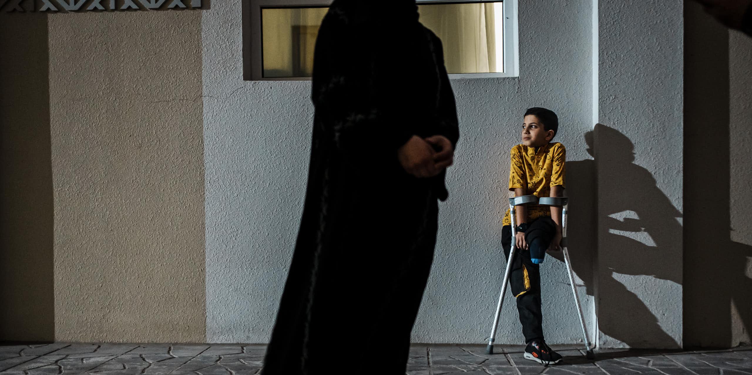 Young boy without a leg uses crutches as he leans against a wall. A faceless adult stands in the foreground.