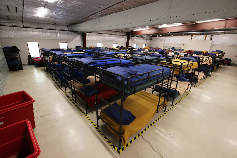 Rows of neatly made bunk beds in a large, brightly lit room.