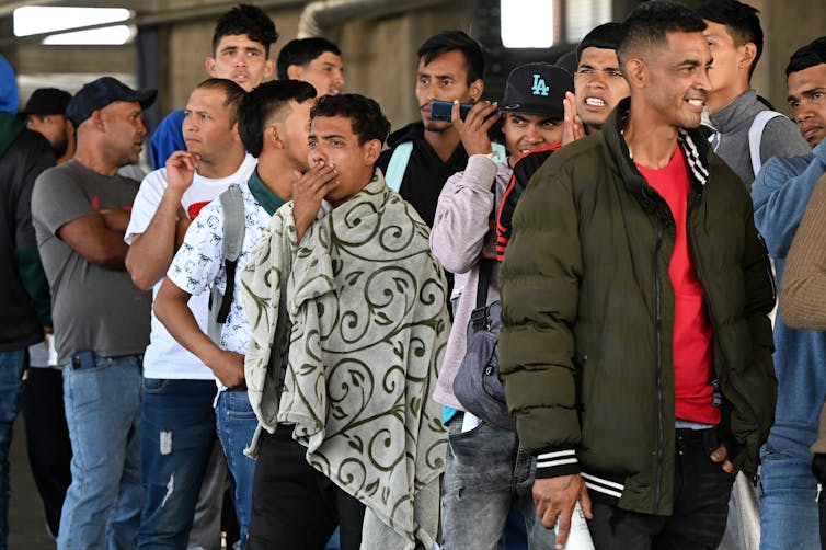 Men stand in line conversing and listening to cell phones.