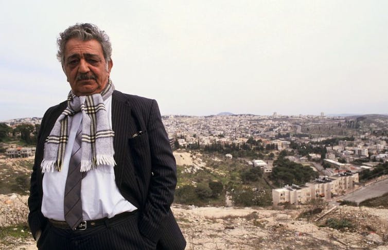 Hombre con bigote vestido con traje y bufanda de pie sobre una colina con vistas a una ciudad.