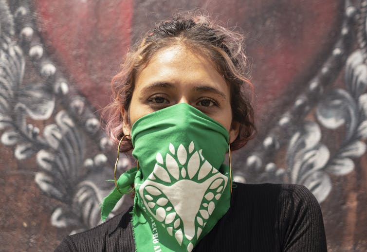 A young Latina woman wears a green headscarf over her nose and mouth