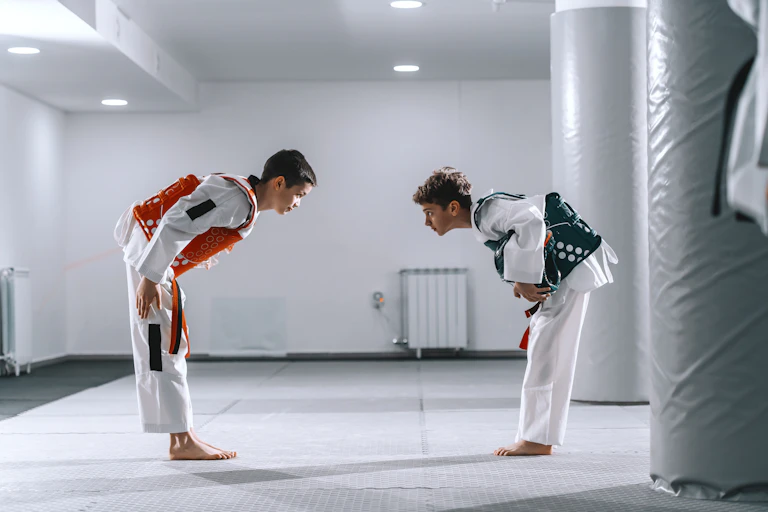 Two boys in white uniforms and colored, puffy vests bow to each other and make eye contact in an all-white room.