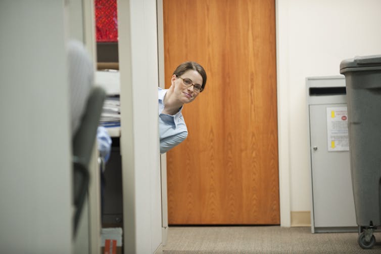 A brunette woman wearing glasses peeks around the wall of an office and looks at the photographer.