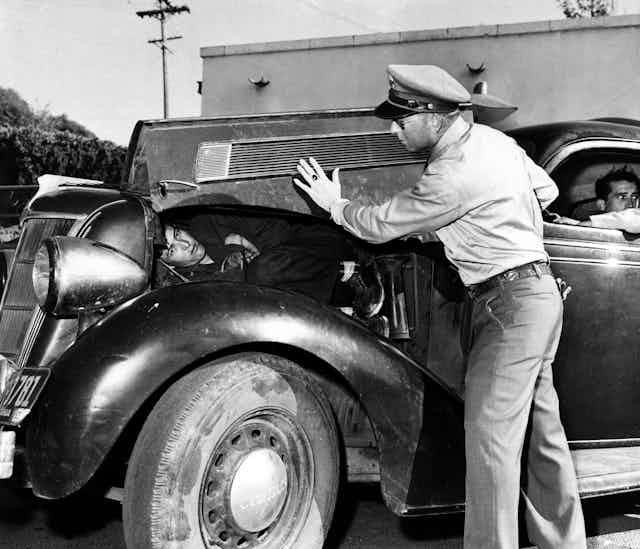 A man with a police hat lifts up the hood of an old car to reveal a man lying there in a black and white photo. 