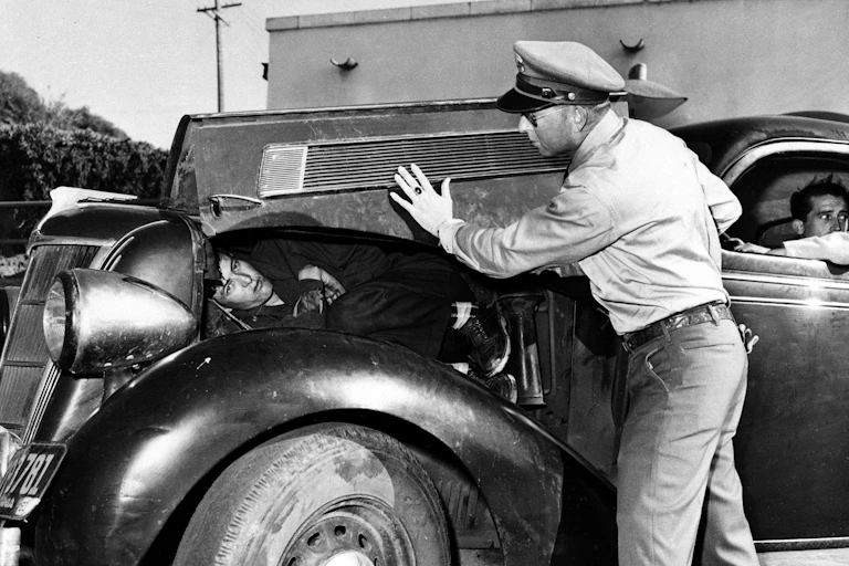 A man with a police hat lifts up the hood of an old car to reveal a man lying there in a black and white photo.