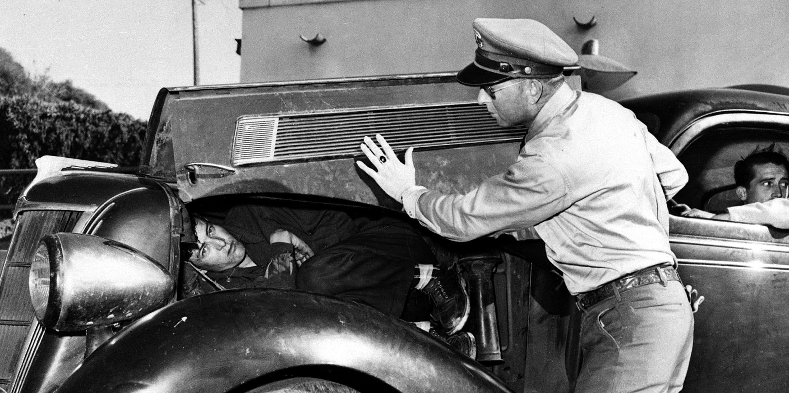 A man with a police hat lifts up the hood of an old car to reveal a man lying there in a black and white photo. 