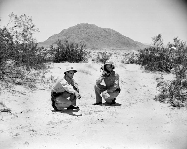 Two men wearing hats crouch on the ground in the desert, with bushes and a mountain around them.