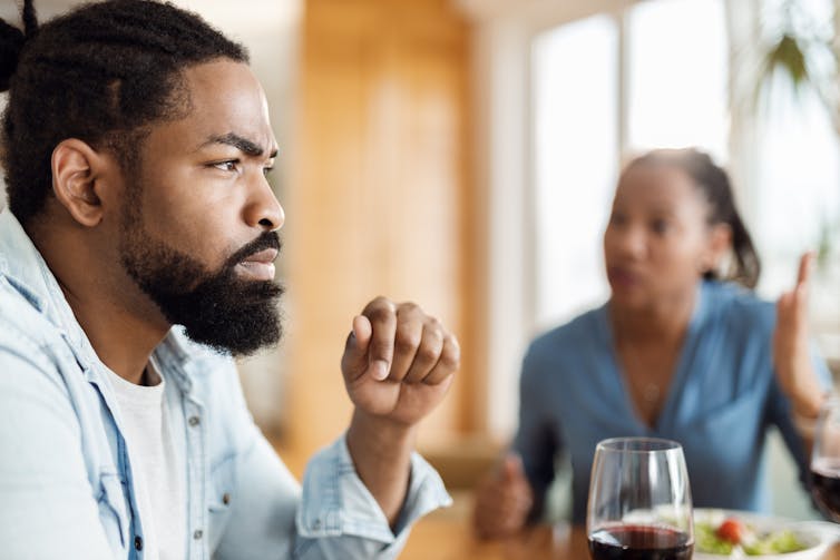 A man with a beard looks into space while a blurry woman sitting at the same table speaks to him.