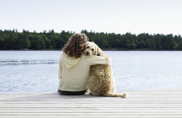 The woman sits on a dock with her arm around her dog, who looks back over her shoulder at the camera.