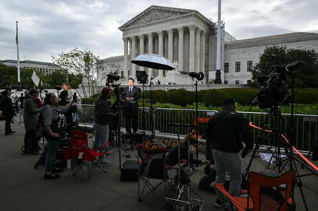 Reporters and camera operators set up in front of a large white, columned building.