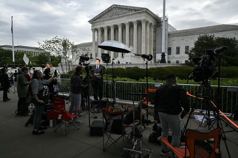 Reporters and camera operators set up in front of a large white, columned building.