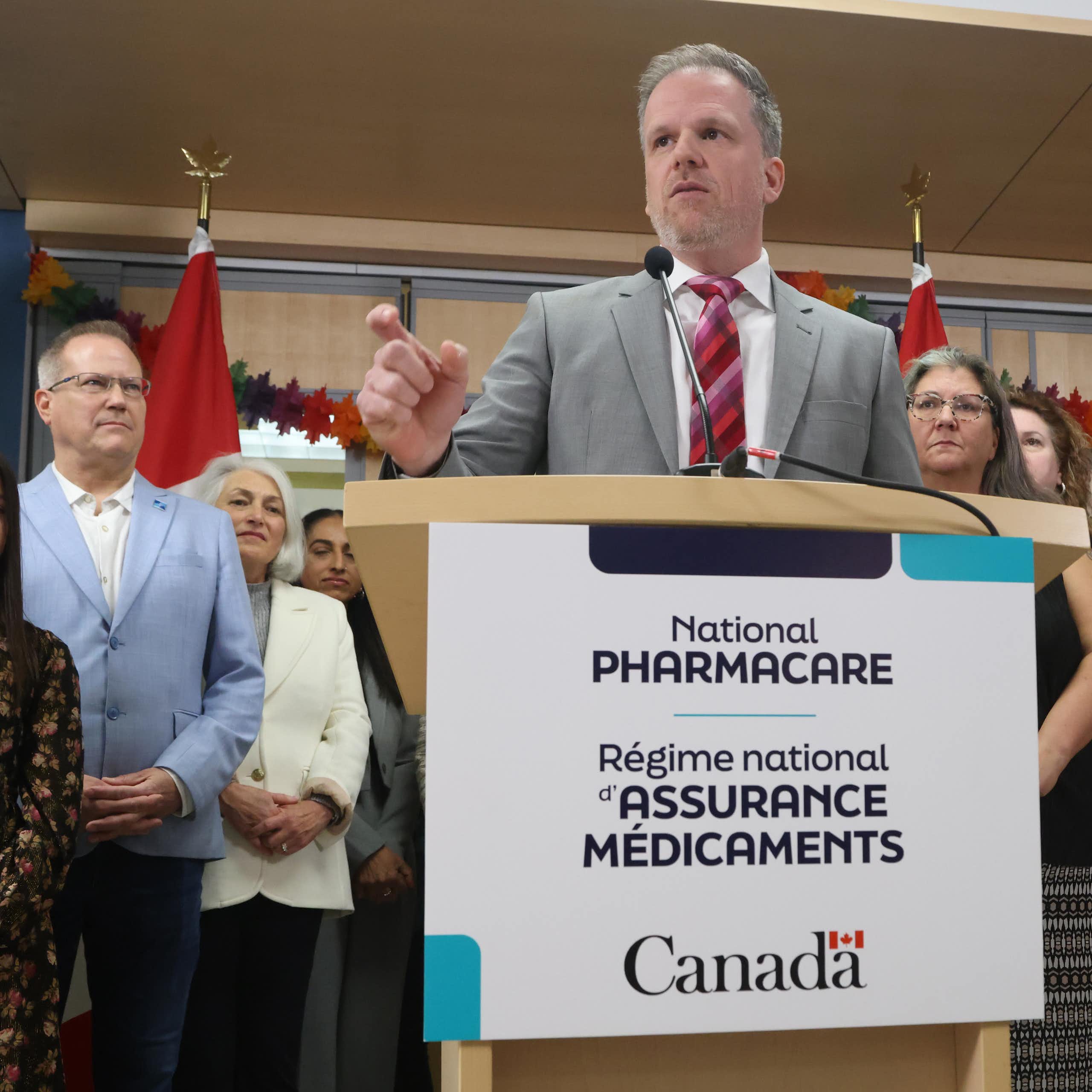 A man in a gray suit standing at a podium marked National Pharmacare