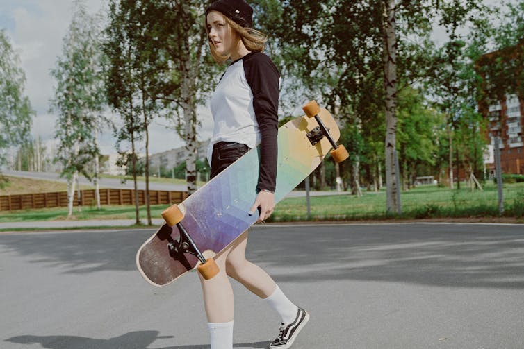 A teenager with long hair carries a skateboard.