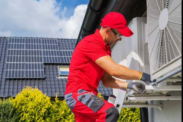 A tradesman in red services a heat pump on the outside of a house.
