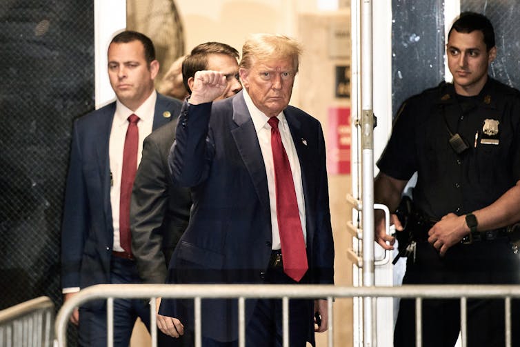 A man in a dark suit and red tie emerging from a building with a police officer near him.