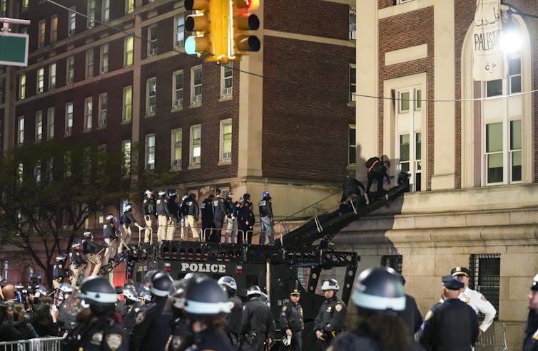 Police in riot gear use a ramp to enter a first floor window in a large apartment building.