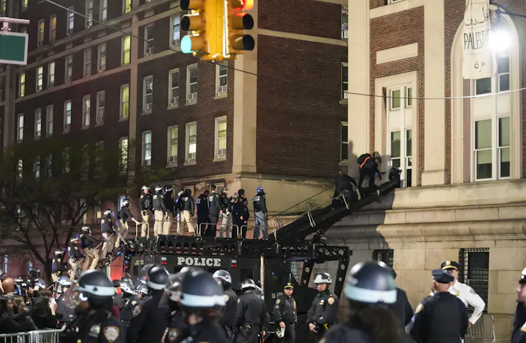 Police in riot gear use a ramp to enter a first floor window in a large apartment building.