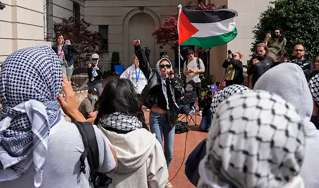 Woman in sunglasses waves a Palestinian red, green, white and black flag surrounded by people wearing keffiyah.