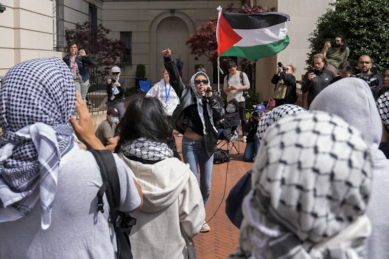 Woman in sunglasses waves a Palestinian red, green, white and black flag surrounded by people wearing keffiyah.