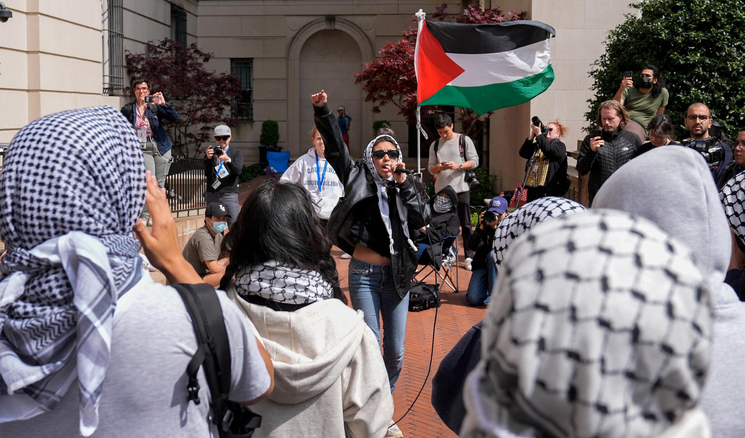 Woman in sunglasses waves a Palestinian red, green, white and black flag surrounded by people wearing keffiyah.