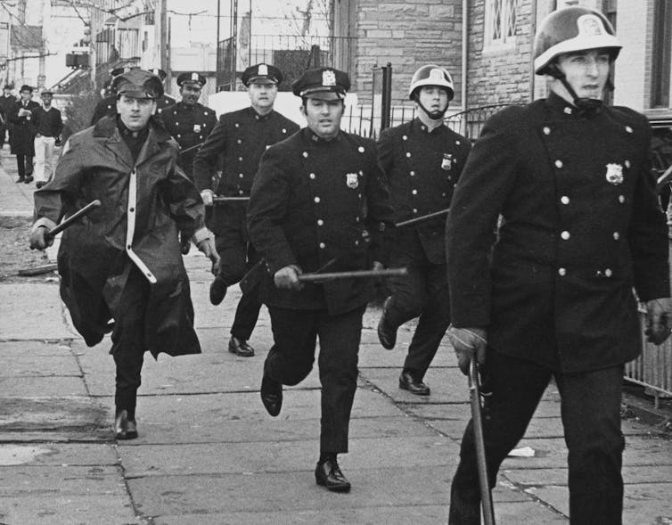Police officers in a black in white vintage photograph wield batons as they move to cut off students.