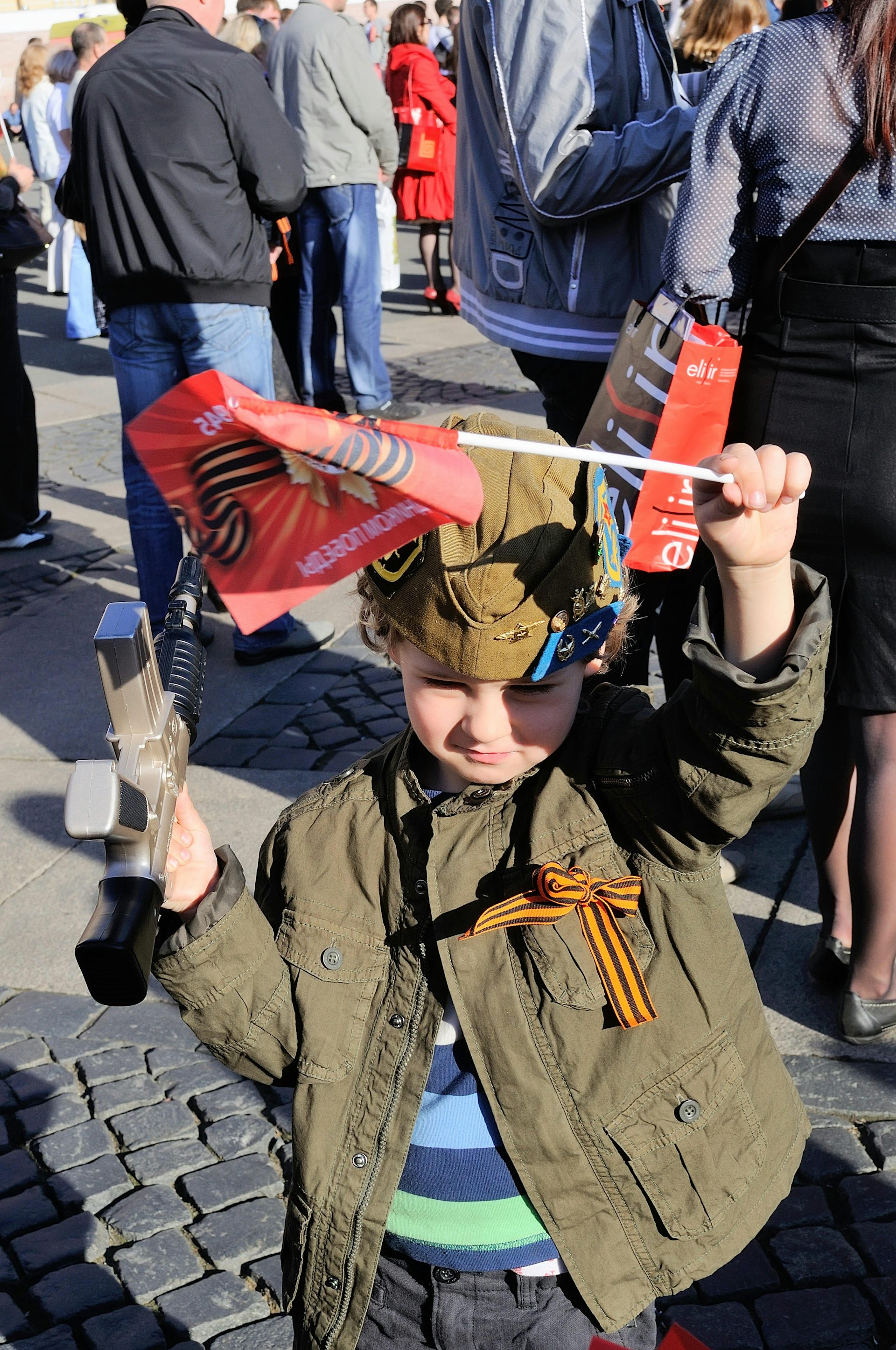 Small boy waving a red flag and wearing a striped ribbon.