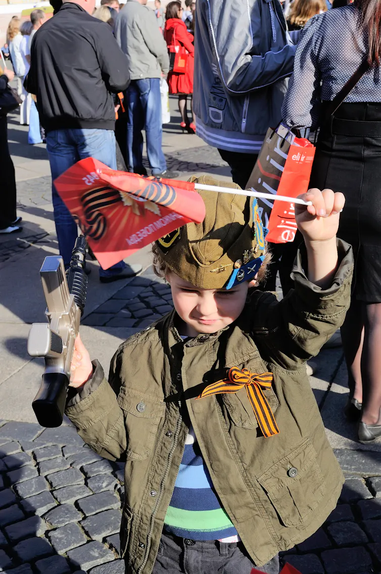Small boy waving a red flag and wearing a striped ribbon.