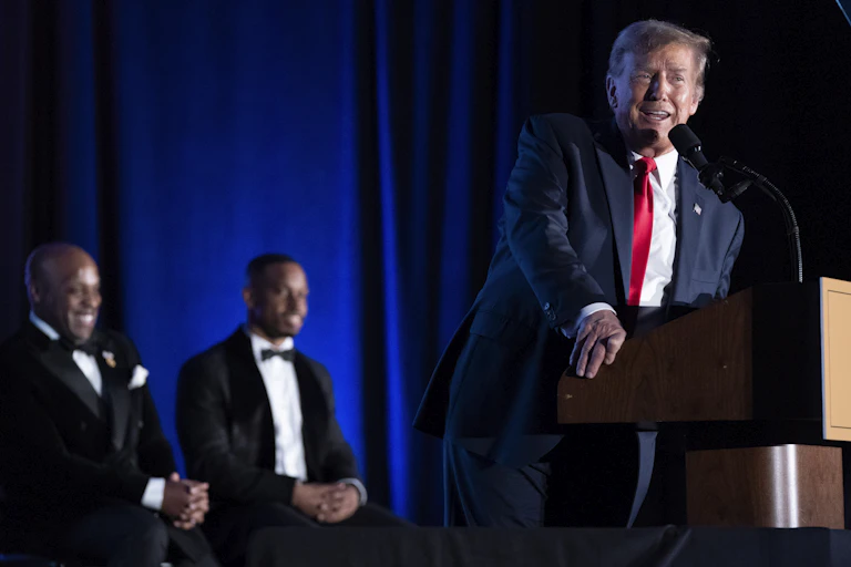 A white man dressed in a business suit is speaking near a lectern two Black men seated in the background are smiling.