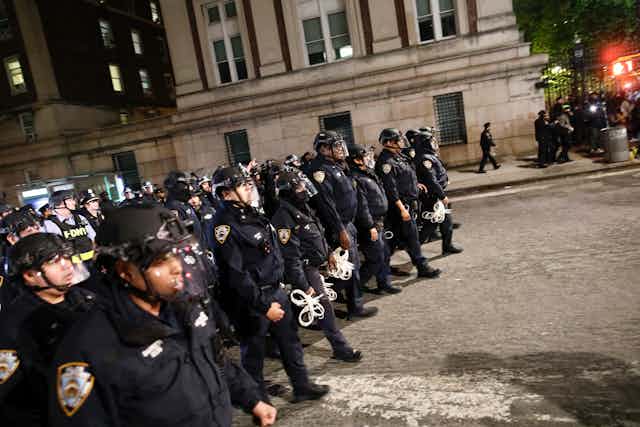 A large crowd of police officers walk down a street with an old building behind them. 