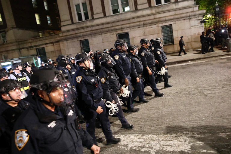 A large crowd of police officers walk down a street with an old building behind them.