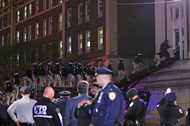 A long line of police officers is seen climbing a black ladder and entering through the window of a building.  Other police officers stand on the ground and watch.