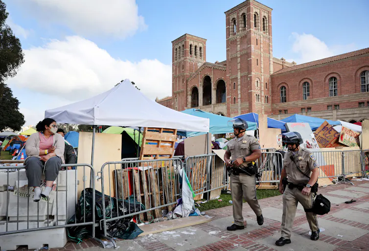Two men in beige uniforms and helmets walk past a woman sitting on a barricade in front of a row of tents.