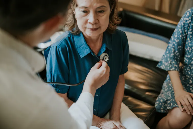 Patient sitting in chair with another person beside them while doctor places stethoscope on their chest