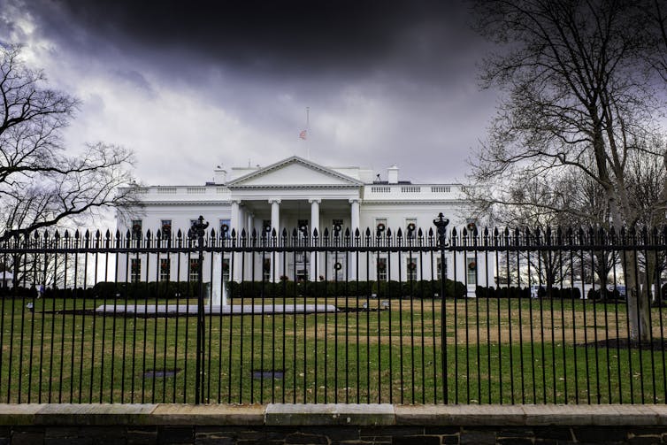A large white mansion with dark clouds over it, behind a fence.