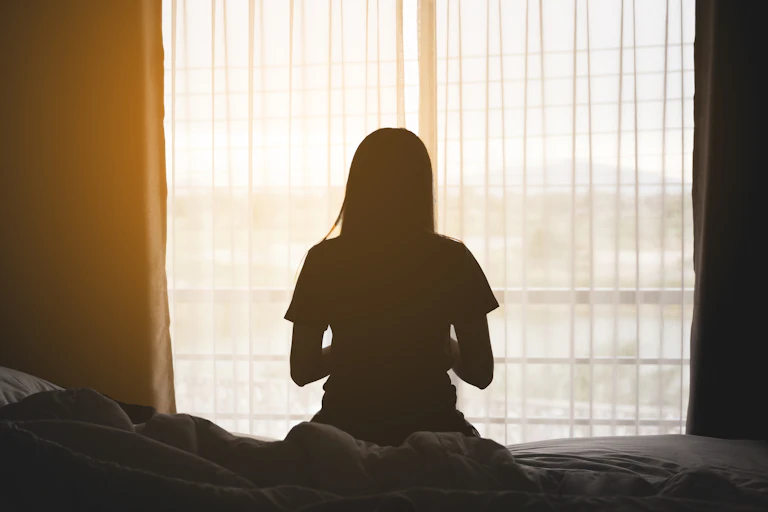 A woman checks her phone while sitting on a bed in front of a window.