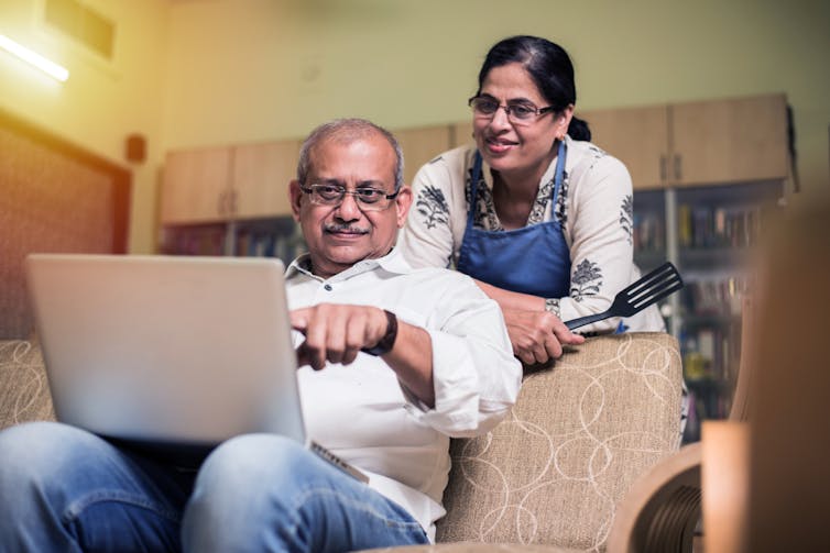 South Asian couple at home, man sitting on sofa pointing at laptop on knees, woman leaning on sofa looking at screen