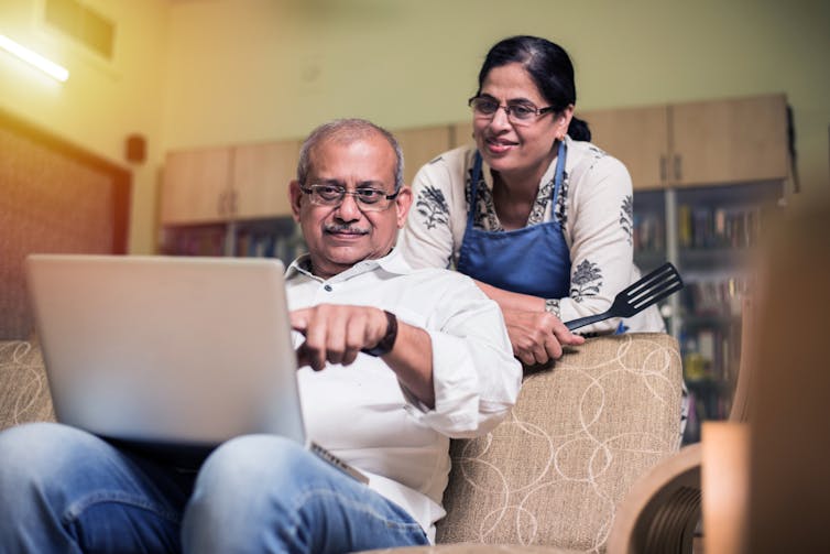 Couple of South Asian descent at home, man sitting on sofa pointing at laptop on knees, woman leaning over sofa looking at screen