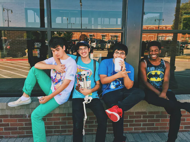 Four young men sit on a ledge in the street. They are hugging and smiling.